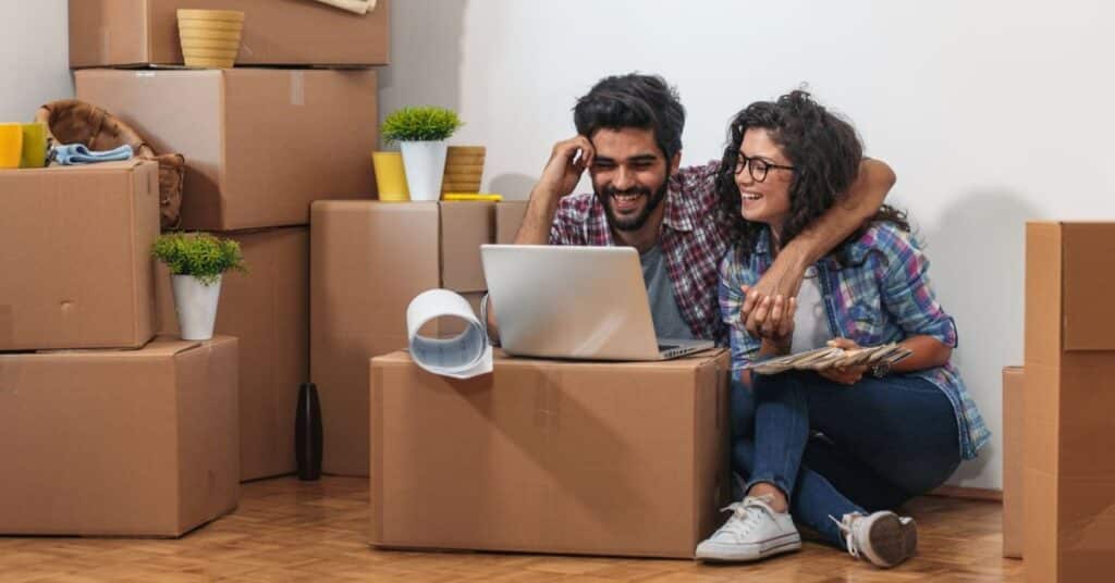 Happy young couple sitting on the floor with their laptop on a cardboard box happy to be moving to a new charity CRM. They are surrounded by moving boxes.