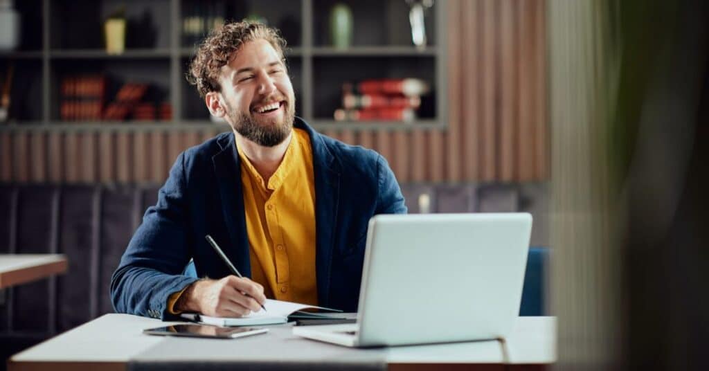 Young smiling man writing notes in agenda and looking at laptop while sitting in cafeteria.